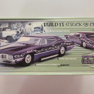 May include: A model car box featuring a purple classic car with flame decals. The box has the text "Build it Stock or Custom!" and "Customs by Flash Customizing Shop". The background shows a vintage garage scene.