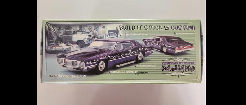 May include: A model car box featuring a purple classic car with flame decals. The box has the text "Build it Stock or Custom!" and "Customs by Flash Customizing Shop". The background shows a vintage garage scene.