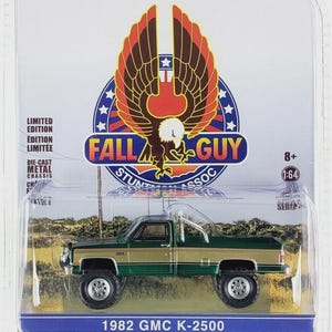 May include: A die-cast metal 1982 GMC K-2500 pickup truck, green and gold in color, in a clear plastic case. The packaging features the text "FALL GUY" and "HOLLYWOOD" with an eagle graphic. The scale is 1:64.