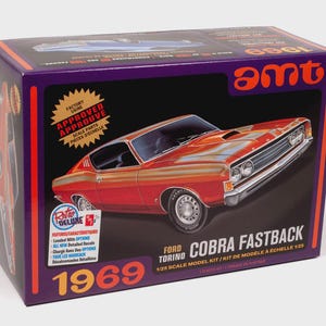 Puede incluir: Caja de modelo a escala de un Ford Torino Cobra Fastback de 1969, de color rojo y naranja. La caja es morada con el logotipo "amt" e incluye texto como "Retro Deluxe" y "Kit de modelo a escala 1:25".