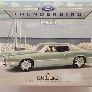 May include: A model kit box featuring a light green 1971 Ford Thunderbird. The box has the Ford logo and the words "THUNDERBIRD 1971". The box also says "MODEL KING 1/25 SCALE MODEL KIT". The background shows a coastal scene with a lighthouse.