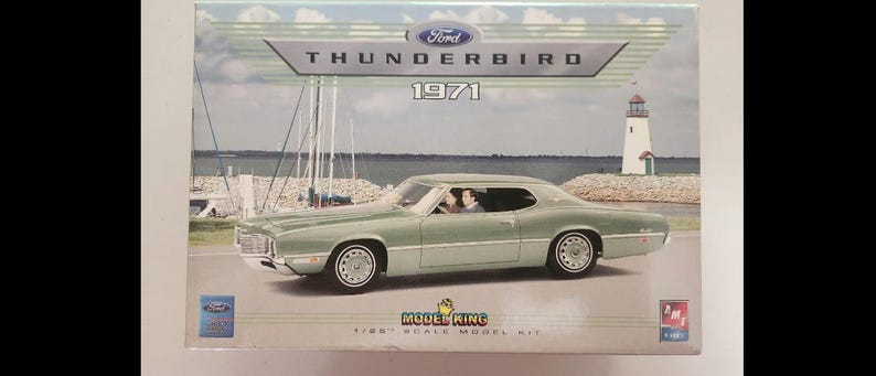 May include: A model kit box featuring a light green 1971 Ford Thunderbird. The box has the Ford logo and the words "THUNDERBIRD 1971". The box also says "MODEL KING 1/25 SCALE MODEL KIT". The background shows a coastal scene with a lighthouse.