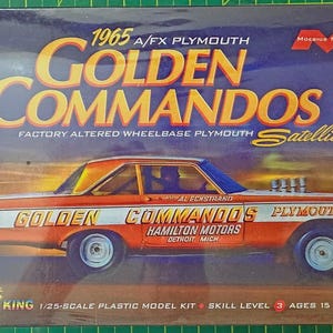 May include: A 1/25-scale plastic model kit box for a 1965 A/FX Plymouth Golden Commandos Satellite. The box features a graphic of an orange and white race car with the text Golden Commandos, Hamilton Motors, Detroit, Mich, and Moebius Models.