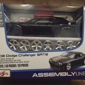 May include: A black 2008 Dodge Challenger SRT8 die-cast metal model kit in a clear plastic box. The kit includes 33 parts and a set of silver wheels. The box also features the Maisto logo and the Assembly Line brand.
