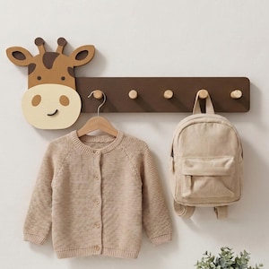 May include: A wooden giraffe-shaped wall hook with a brown board and five pegs. A beige cardigan hangs from a hook, and a small backpack is attached. A potted plant sits below.