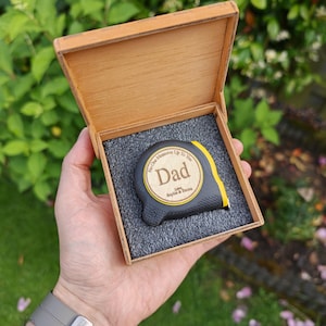 Peut inclure: Un mètre ruban noir et jaune avec une incrustation circulaire en bois gravée du texte No One Measures Up To You Dad Love, Sophie & Emma, présenté dans une boîte en bois carrée avec un insert en mousse gris foncé pour le rangement.