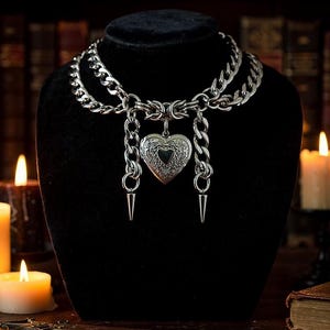 May include: A silver-toned necklace with a heart-shaped locket pendant and two dangling spike charms. The necklace features a double-layered chain design and is displayed on a black mannequin head. The background includes candles and a bookshelf.