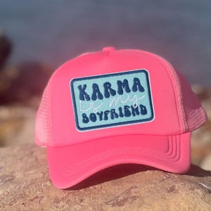 May include: A bright pink trucker hat with a mesh back and a curved brim. The front features a rectangular patch with the words "Karma is my Boyfriend" in white and blue lettering.