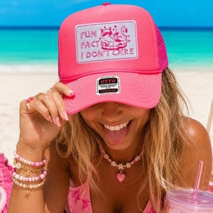 May include: A bright neon pink trucker hat featuring a rectangular patch with a graphic of a raccoon and a flamingo in a pool float, accompanied by the text "FUN FACT I DON'T CARE". The hat includes a black and white Otto brand sticker on the brim.