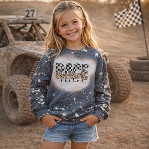 May include: A young girl wearing a gray sweatshirt with the words "RACE sister" in a checkered and leopard print design. The sweatshirt has white paint splatters. She is wearing denim shorts and smiling.