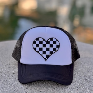 May include: A black and white checkered heart design embroidered on a white trucker hat with a black mesh back.
