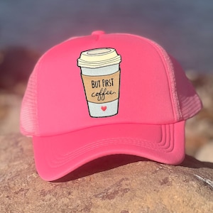 Puede incluir: Gorra de camionero rosa brillante con un diseño de taza de café. El diseño presenta una taza de café con el texto "BUT FIRST coffee" y un pequeño corazón rojo. La gorra tiene una parte trasera de malla y una visera curva.