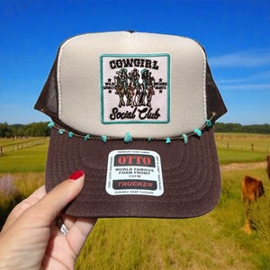 May include: A brown and tan trucker hat with a mesh back and a patch that reads "COWGIRL Social Club." The hat has turquoise-colored beads along the brim. The hat is made of foam and has a snap closure.