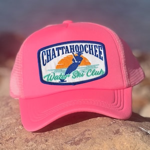 May include: A bright pink trucker hat with a mesh back and a curved brim. The hat features a rectangular patch with the words "Chattahoochee Water Ski Club" and a graphic of a skier in blue and orange. The hat is made of a durable material.