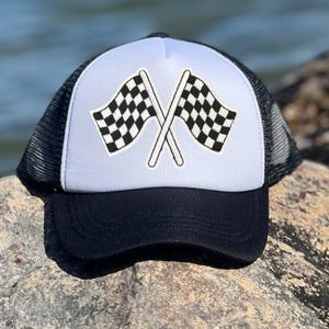 以下が含まれることがあります： 白のフロントパネルに2つの交差したチェッカーフラッグが描かれた、白と黒のトラッカーハット。 黒いツバと黒いメッシュの背面を備えた帽子で、モータースポーツ愛好家に適しています。