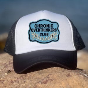 May include: A black and white trucker hat with a white front panel and a black mesh back. The hat features a patch with the words "CHRONIC OVERTHINKERS CLUB" in black text on a light blue background, with floral accents.