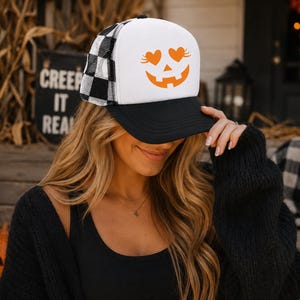 Peut inclure: Une casquette de type trucker avec un panneau avant blanc orné d'un motif orange représentant un visage de citrouille d'Halloween, avec des yeux en forme de cœur et un sourire. Elle possède une visière noire et un filet arrière à carreaux noirs et blancs.