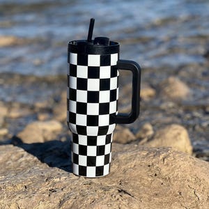 Puede incluir: Un vaso de doble pared negro y blanco a cuadros con un asa negra y una pajita negra. El vaso está sentado en una roca.