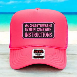 May include: A vibrant pink trucker hat with a mesh back and a curved peak. The front displays a black patch with the text "YOU COULDN'T HANDLE ME EVEN IF I CAME WITH INSTRUCTIONS" in pink. A small white tag is also visible.