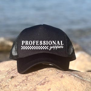Könnte beinhalten: Schwarze Trucker-Mütze mit den weißen Worten "PROFESSIONAL yapper" und einem Schachbrettmuster. Die Mütze hat einen Mesh-Rücken und einen gebogenen Schirm, der auf einem Felsen mit verschwommenem Ozeanhintergrund ruht.