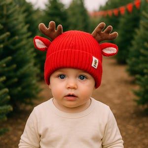 Pode incluir: Um gorro de malha canelada vermelho brilhante com chifres de rena de pelúcia castanhos e orelhas forradas a branco nas laterais. Uma pequena etiqueta retangular branca com um logótipo de brasão dourado está cosida na dobra frontal.