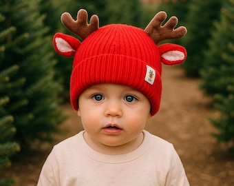 Puede incluir: Un gorro de punto acanalado de color rojo brillante con astas de reno de felpa marrón y orejas forradas en blanco a los lados. El gorro incluye una pequeña etiqueta blanca rectangular con un logotipo gráfico en el dobladillo, sobre una camisa crema.