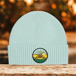 Puede incluir: Un gorro de punto azul claro con un parche circular. El parche presenta un sol amarillo, cielo azul, árboles verdes y las palabras "SEEK ADVENTURE". El gorro está hecho de punto acanalado.