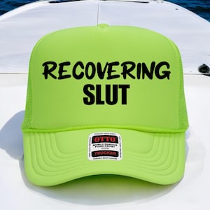 May include: A neon green trucker hat with the words "RECOVERING SLUT" in bold black letters. The hat has a mesh back and a foam front. A small white sticker with black text is on the front.