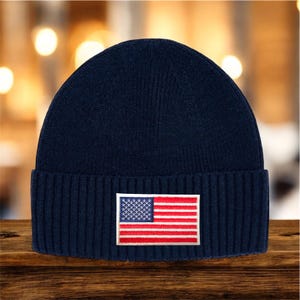 May include: A navy blue knit beanie with a ribbed cuff. The beanie features a rectangular patch with the American flag design, showcasing red and white stripes and a field of white stars on a blue background.