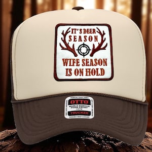 May include: A beige and brown trucker hat featuring a square patch with a dark red border. The patch displays brown deer antlers, a black crosshair symbol, and the text IT'S DEER SEASON WIFE SEASON IS ON HOLD. An OTTO brand sticker is on the brim.