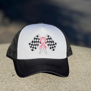 May include: A white and black trucker hat with a pink ribbon and checkered flags embroidered on the front.