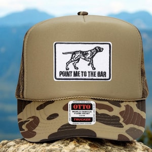 May include: A tan trucker hat with a brown mesh back and a camouflage pattern on the brim. The hat features a rectangular patch with a dog illustration and the text "POINT ME TO THE BAR".