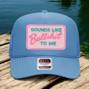 May include: A light blue trucker hat with a mesh back and a curved brim. The front features a pink rectangle with the words "SOUNDS LIKE Bullshit TO ME" in green and pink lettering. The hat has a snap closure.