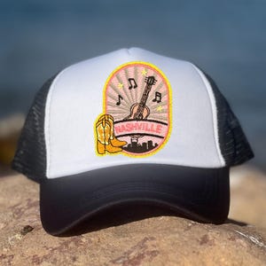 Puede incluir: Gorra de camionero blanca y negra con un parche bordado. El parche presenta una guitarra, notas musicales, una bota de vaquero y la palabra "Nashville". El diseño es rosa y amarillo, evocando la música country.
