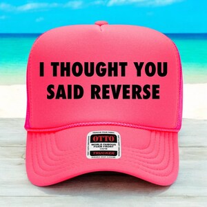 May include: A vibrant pink trucker hat featuring the black text "I THOUGHT YOU SAID REVERSE". The hat has a mesh back and a curved peak. The hat has an "OTTO" brand label.