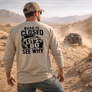 May include: A light tan long-sleeved shirt with the text "ROAD IS CLOSED LET'S GO SEE WHY" and a jeep graphic. A person wearing a camouflage baseball cap and sunglasses is seen from the back, with a jeep driving in the background.