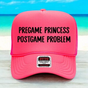 May include: A bright pink trucker hat with the text "PREGAME PRINCESS POSTGAME PROBLEM" in black. The hat has a mesh back and a curved brim. The hat also has a small white tag with the brand name "OTTO".