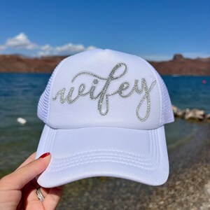 Peut inclure: Casquette de camionneur blanche avec le mot "wifey" en strass argentés. Le chapeau a un panneau avant blanc et un dos en filet blanc. La visière est incurvée. Le chapeau est tenu par une main.