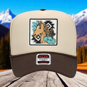 May include: A trucker hat with a brown brim and a tan crown. The front features a square patch with a horse illustration, turquoise accents, and a black and white geometric design. The hat has a brown mesh back and a snap closure.