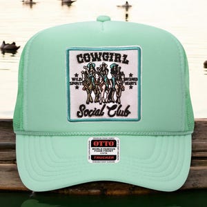 May include: A mint green trucker hat with a mesh back and a foam front. The front patch reads "COWGIRL Social Club" with an illustration of cowgirls on horses. The hat also features a small white tag with the brand name "OTTO".
