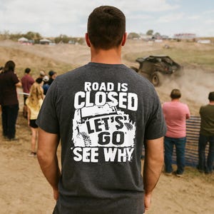 May include: A charcoal grey t-shirt with the white text "ROAD IS CLOSED LET'S GO SEE WHY". The graphic features an off-road vehicle. The person is outdoors, observing an off-road event with other spectators.