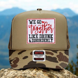 May include: A trucker hat with a tan front panel and a camouflage patterned brim. The hat features a patch with the text "WE GO Together LIKE DRUNK & DISORDERLY" in red and white. The hat has a mesh back and a snap closure.