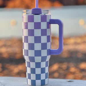 Puede incluir: Un vaso grande con un patrón de cuadros morados y blancos. El vaso tiene un asa, tapa y pajita moradas a juego. La taza está diseñada para mantener las bebidas frías o calientes. El vaso está sobre una superficie blanca.