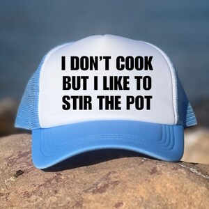 Pode incluir: Boné de caminhoneiro branco e azul claro com o texto "I DON'T COOK BUT I LIKE TO STIR THE POT" em preto. O boné tem um painel frontal branco, uma aba azul claro e uma parte traseira de malha.