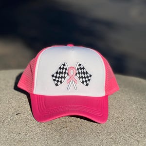May include: A pink and white trucker hat with a checkered flag and pink ribbon design. The hat has a mesh back and a pink brim.