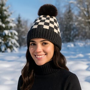 Pode incluir: Um gorro de inverno com um padrão xadrez preto e branco e um pompom castanho escuro. O gorro é usado por uma pessoa num cenário exterior com neve. A pessoa está a usar uma camisola de gola alta preta.