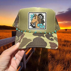 May include: A green trucker hat with a camouflage pattern on the brim and a square patch featuring a horse illustration. The hat is adorned with turquoise-colored beads along the brim, adding a decorative touch. The hat is held by a person.