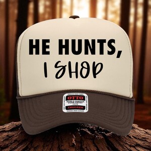 May include: A trucker hat with a brown brim and mesh back. The front panel is beige with the text "HE HUNTS, I SHOP" in black. The hat has a snap closure and a small label that reads "OTTO".