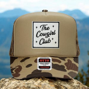 May include: A trucker hat with a tan front and a camouflage patterned brim. The hat features a white square patch with the text "The Cowgirl Club" in black script. The hat has a mesh back and a snap closure.