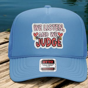 May include: A light blue trucker hat with a mesh back. The front of the hat features a patch with the words "WE LISTEN, and WE JUDGE" in a playful font. The patch includes heart and flower graphics.